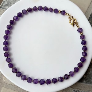 May include: A purple amethyst gemstone necklace with a gold clasp. The necklace is made of faceted beads and is approximately 18 inches long.