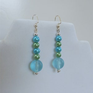 May include: A pair of dangle earrings with a light blue and green color scheme. The earrings are made with round beads and have a silver hook.