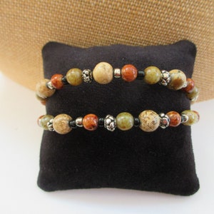 May include: Two bracelets made with brown, green, and black beads with silver accents. The bracelets are displayed on a black velvet cushion.