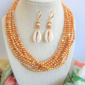 May include: A multi-strand necklace and matching earrings made of peach-coloured freshwater pearls. The earrings feature a small white seashell charm.