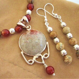 K&ouml;nnte beinhalten: Ein Schmuckset mit Halskette und Ohrringen. Die Halskette hat rote und beige Perlen mit einem silbernen Anh&auml;nger. Die Ohrringe haben beige Perlen und silberne Akzente. Der Anh&auml;nger ist ein gro&szlig;er, runder Stein mit einem silbernen Rahmen.
