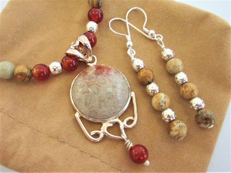K&ouml;nnte beinhalten: Ein Schmuckset mit Halskette und Ohrringen. Die Halskette hat rote und beige Perlen mit einem silbernen Anh&auml;nger. Die Ohrringe haben beige Perlen und silberne Akzente. Der Anh&auml;nger ist ein gro&szlig;er, runder Stein mit einem silbernen Rahmen.