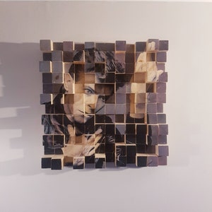 Puede incluir: Una pieza de arte de mosaico de madera que muestra una foto en blanco y negro de un hombre con cabello corto y oscuro. La foto está dividida en pequeños cuadrados de madera, creando un efecto pixelado.