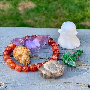 May include: A collection of Halloween-themed crystal carvings, including a bat, skull, ghost, and crescent moon. The crystals are arranged on a grey wooden surface with a carnelian bead bracelet.