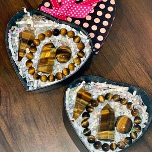 May include: Two heart-shaped boxes filled with tiger's eye gemstone jewelry. Each box contains a bracelet, a pendant, and a sphere. The boxes are lined with white tissue paper and have a black and white polka dot pattern.