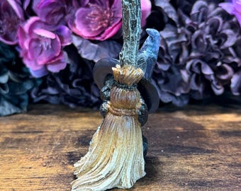 May include: A small figurine of a witch riding a broom. The witch is wearing a black hat and holding onto a wooden broom with a straw broom head. A blue bird is perched on the broom handle. The background features purple flowers.