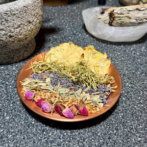 May include: A copper-colored dish filled with dried herbs, flowers, and pineapple slices. The arrangement includes dried lavender, rosemary, and rosebuds. A mortar and pestle are visible in the background, suggesting a natural wellness theme.