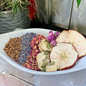 Könnte beinhalten: Eine weiße Schüssel gefüllt mit getrockneten Früchten, Blumen und Gewürzen. Die Schüssel enthält getrocknete Apfelscheiben, Lavendel, Rosenblüten, Zimtstangen und Kardamomkapseln.