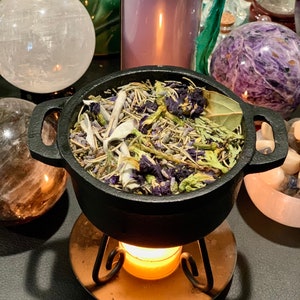 Può includere: Un calderone di ghisa nero pieno di un mix di erbe e fiori secchi, posto su un supporto in metallo con una candela accesa sotto. Il calderone è circondato da cristalli e altri oggetti decorativi.