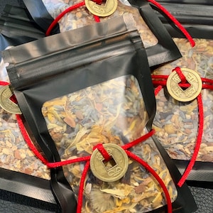 Könnte beinhalten: Mehrere transparente Plastiktüten gefüllt mit getrockneten Kräutern und Blumen. Jede Tüte ist mit einem schwarzen Reißverschluss verschlossen und mit einer roten Kordel gebunden. An jeder Tüte ist eine goldfarbene Münze mit einem quadratischen Loch befestigt.