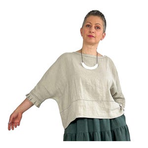 May include: A beige linen top with a wide boat neckline, three-quarter length sleeves featuring ruffled cuffs, and a horizontal row of dark decorative stitching across the midsection, paired with a dark green tiered skirt and a white crescent necklace.