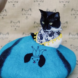 May include: A black and white cat wearing a patterned bandana with yellow trim sits in a teal blue felt cat bed with a black and white embroidered dog face with a crown on top.