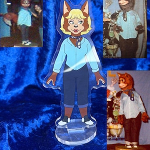 May include: A clear acrylic standee featuring a cartoon-style anthropomorphic fox character with blonde hair, wearing a light blue button-down shirt, dark blue pants, and a patterned neck scarf, standing on a circular transparent base.
