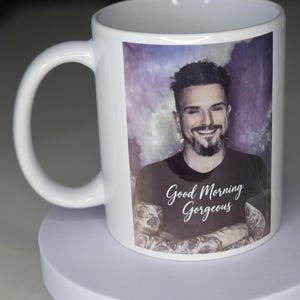 May include: A white ceramic mug featuring a printed image of a smiling person with a goatee and arm tattoos. The person wears a black t-shirt with the white text Good Morning Gorgeous printed on the front. The background of the image is purple.