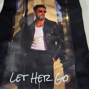 Pode incluir: Uma t-shirt preta com uma estampa gráfica de uma pessoa vestindo uma regata branca, uma jaqueta de couro preta e óculos de sol, em pé perto de uma coluna de pedra. O texto "LET HER GO" está impresso em branco na parte inferior da imagem.