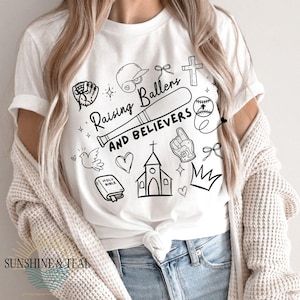 May include: White t-shirt with a black graphic design featuring a baseball bat, baseball, cross, and the text "Raising Ballers AND BELIEVERS." Additional illustrations include a baseball glove, helmet, dove, heart, church, and crown.