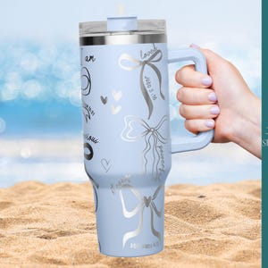 Puede incluir: Un vaso t&eacute;rmico azul claro con asa, decorado con lazos plateados, corazones y escritura. El vaso tiene una tapa transparente y se sostiene en una playa de arena con el oc&eacute;ano al fondo. El texto incluye "amado" y "poderoso".