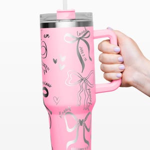 Puede incluir: Un vaso t&eacute;rmico rosa con asa, tapa y pajita. El vaso est&aacute; decorado con lazos plateados, corazones y texto inspirador, incluyendo "I am", "loved" y "powerful". El asa y la tapa son del mismo color rosa que el cuerpo del vaso.