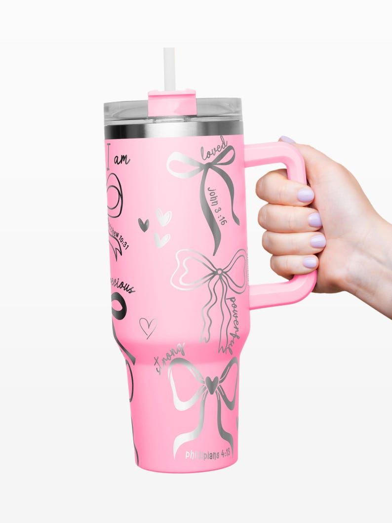 Puede incluir: Un vaso t&eacute;rmico rosa con asa, tapa y pajita. El vaso est&aacute; decorado con lazos plateados, corazones y texto inspirador, incluyendo "I am", "loved" y "powerful". El asa y la tapa son del mismo color rosa que el cuerpo del vaso.