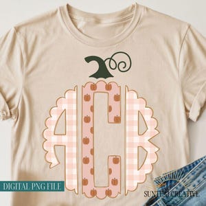 May include: A beige t-shirt featuring a large graphic of a scalloped monogram with the letters A, C, and B. The outer sections have a light pink gingham pattern, while the center section is pink with small orange pumpkin icons and a dark green stem.