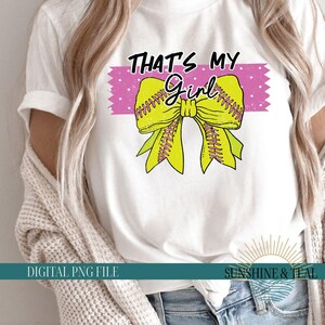 May include: White t-shirt with a pink and white ribbon design. The ribbon is made of yellow softball stitching. The text "That's My Girl" is printed on the shirt.