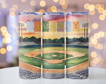 May include: Three stainless steel tumblers with a colorful baseball field scene and the word "GAMEDAY" in colorful letters. The tumblers are on a white surface with a blurred background of lights.