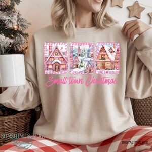 Puede incluir: Sudadera beige con un gráfico rosa de tres casas de acuarela en una escena invernal. El texto "Small Town Christmas" está en rosa debajo de las casas. Las palabras "Sunshine & Teal" están en la parte inferior.
