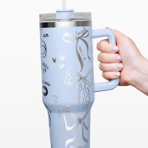 Puede incluir: Un vaso aislado azul claro con asa y pajita. El vaso presenta dise&ntilde;os de lazo plateado y texto inspirador, incluyendo "I am", "loved" y vers&iacute;culos b&iacute;blicos. El vaso es sostenido por una persona.