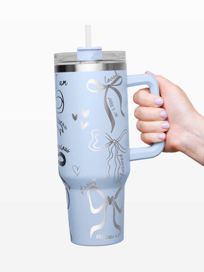 Puede incluir: Un vaso aislado azul claro con asa y pajita. El vaso presenta dise&ntilde;os de lazo plateado y texto inspirador, incluyendo "I am", "loved" y vers&iacute;culos b&iacute;blicos. El vaso es sostenido por una persona.
