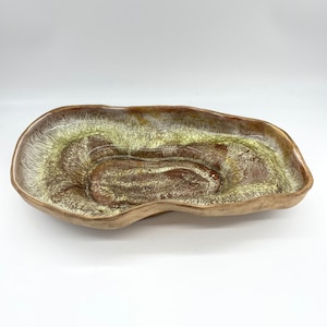 May include: A large, free-form ceramic serving dish with a brown and green glaze. The dish has a deep, irregular shape and a glossy finish.