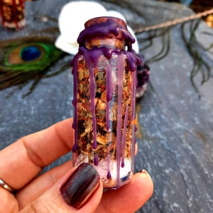 May include: A small glass bottle filled with dried herbs and crystals, sealed with a purple wax top. The bottle is held in a hand.