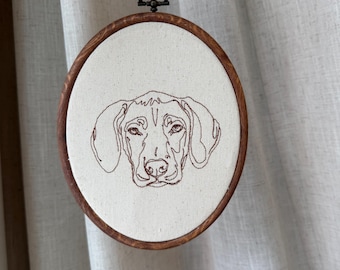 May include: An embroidered dog portrait in a wooden hoop. The dog's face is rendered in brown thread on a cream-colored fabric. The hoop is round, with a dark brown finish, and has a metal hanging loop at the top.