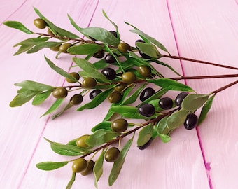 May include: A sprig of olive branches with green leaves and black and green olives on a pink wooden surface.