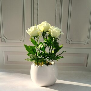 May include: A white ceramic egg-shaped planter filled with white roses and green foliage. The planter has a scalloped rim and sits on a white surface. The background is a white wall with decorative molding.