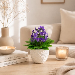 Può includere: Un tavolino rotondo in legno mostra un vaso bianco strutturato con una pianta di violette. Una candela accesa in un portacandele di vetro e due libri sono anche sul tavolo. Un vaso beige con fiori secchi è sullo sfondo.