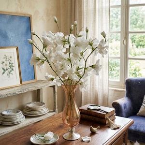 May include: A bouquet of white flowers in a peach-colored glass vase on a wooden table. The table also holds books, a magnifying glass, and decorative plates. A blue armchair and a window with a view are in the background.