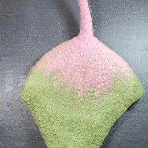 May include: A handmade felted hat in a unique shape, featuring a color gradient from light green to pink. The hat has a curved, elongated pink stem. The felted texture is visible, showcasing the craftsmanship of the piece.