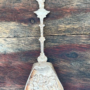 May include: A silver-coloured metal souvenir spoon featuring a sculpted figure of Manneken Pis at the top of the handle. The wide, shovel-shaped bowl displays a detailed embossed scene of three figures seated at a table within an interior setting.