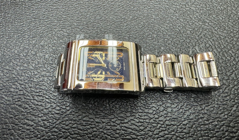 K&ouml;nnte beinhalten: Eine silberne und goldene Herrenarmbanduhr mit quadratischem Zifferblatt und einem Metallband.
