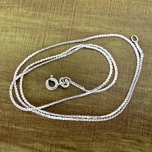 May include: A silver-toned chain necklace featuring a delicate, textured link design. The necklace includes a circular spring ring clasp and a small jump ring at the opposite end, showcasing a polished metallic finish throughout the entire piece.