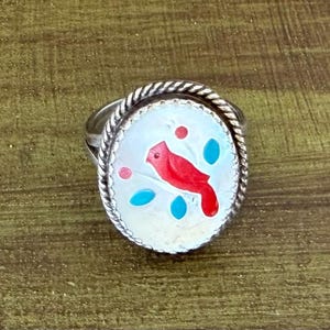 May include: Oval silver ring featuring a red cardinal bird design on a white background. The bird is surrounded by turquoise leaves and red dots. The ring has a twisted rope-like border.