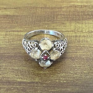 May include: A silver ring with a floral design, featuring five light-colored gemstones and a small red gemstone at the center. The ring has an ornate, openwork band.