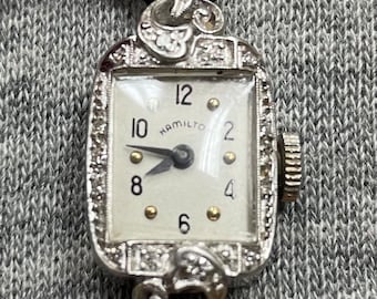 Könnte beinhalten: Eine vintage silberne Armbanduhr mit weißem Zifferblatt und schwarzen Zeigern. Die Uhr hat ein quadratisches Zifferblatt mit Diamantenbesatz am Rand. Das Zifferblatt der Uhr hat schwarze römische Ziffern für die Stunden und das Wort "Hamilton" in Schwarz gedruckt.
