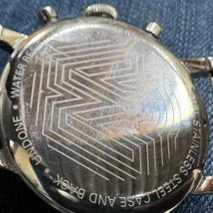 May include: Close-up of the back of a stainless steel watch. The watch back features an etched geometric design and the words "WATER RESIST" and "STAINLESS STEEL CASE AND BACK".
