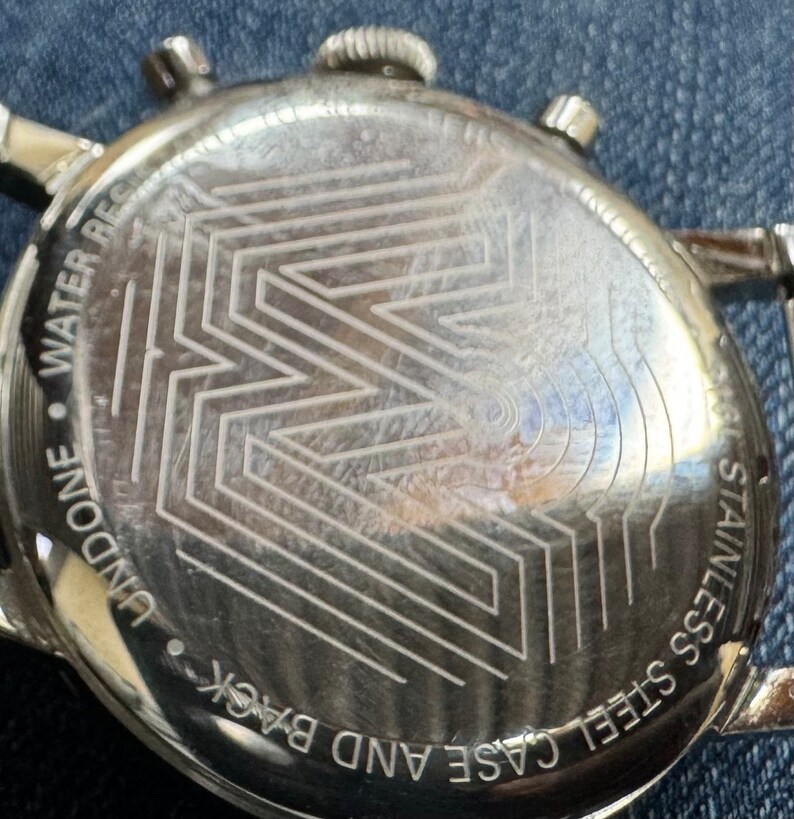May include: Close-up of the back of a stainless steel watch. The watch back features an etched geometric design and the words "WATER RESIST" and "STAINLESS STEEL CASE AND BACK".