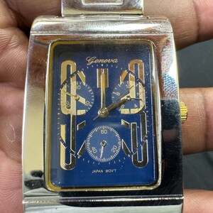 K&ouml;nnte beinhalten: Eine silberne und goldene Chronographen-Armbanduhr mit blauem Zifferblatt und dem Wort "Geneva" auf dem Zifferblatt. Die Uhr hat drei Hilfszifferbl&auml;tter und den Text "JAPAN MOV'T" auf dem Zifferblatt.