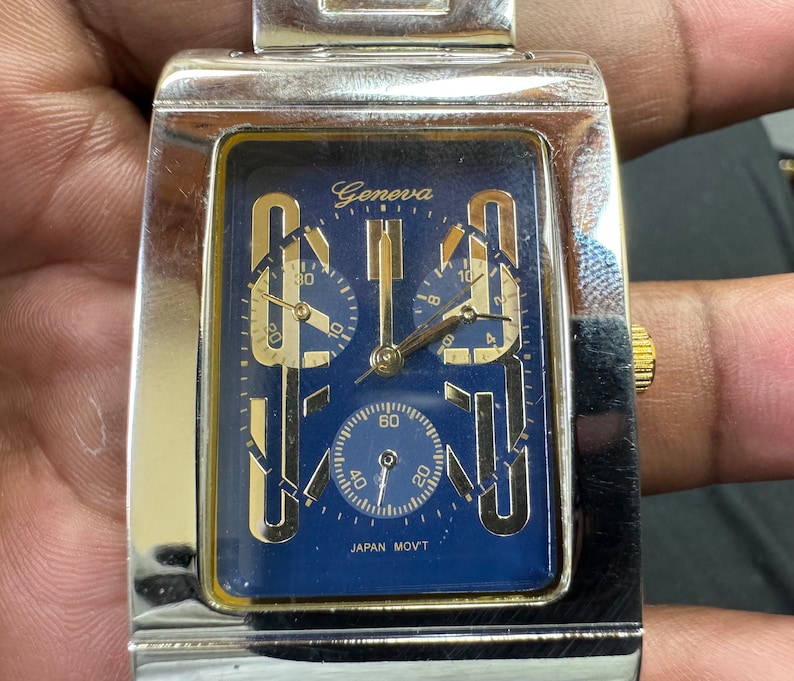 K&ouml;nnte beinhalten: Eine silberne und goldene Chronographen-Armbanduhr mit blauem Zifferblatt und dem Wort "Geneva" auf dem Zifferblatt. Die Uhr hat drei Hilfszifferbl&auml;tter und den Text "JAPAN MOV'T" auf dem Zifferblatt.