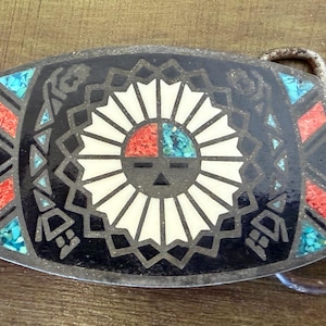 May include: A rectangular metal belt buckle featuring a central sun face motif with inlaid red and turquoise stone segments. The design is surrounded by a black background with geometric patterns of red and turquoise stone inlays on the sides.