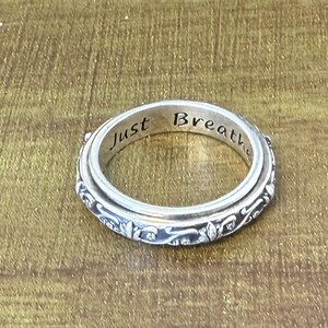 May include: A silver-toned spinner ring featuring an intricate floral scrollwork pattern on the outer band and the engraved text Just Breathe visible on the inner surface of the ring. The design consists of a rotating outer band over a fixed base.