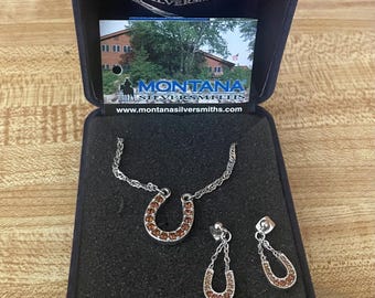 May include: A silver horseshoe necklace and earrings set with brown crystal accents. The jewelry is displayed in a blue velvet box with the text "Montana Silversmiths" on the lid.